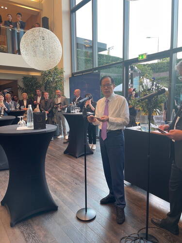 Älterer Mann im Anzug mit lila Krawatte hält eine Rede am Mikrofon in einem modernen, lichtdurchfluteten Veranstaltungsraum, umgeben von aufmerksam zuhörenden Gästen an Stehtischen.
