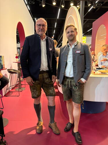 Zwei Männer in Trachtenjacken, Lederhosen und Haferlschuhen stehen lächelnd auf einem modernen Messestand mit pinkem Boden und beleuchteten, bogenförmigen Elementen im Hintergrund