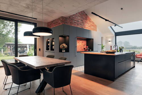 Offene Designküche mit schwarzer Kochinsel, gemauerter Backsteinwand, großem Holzesstisch und bodentiefen Fenstern mit Blick in den Garten