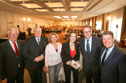 Sechs lächelnde Rednerinnen und Redner posieren im Vordergrund eines voll besetzten Konferenzsaals vor dem Publikum.