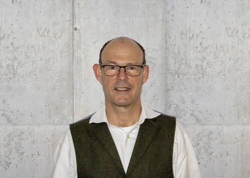 Mittelalterlicher Mann mit Brille, weißem Hemd und dunkelgrüner Weste steht lächelnd vor einer grauen Betonwand