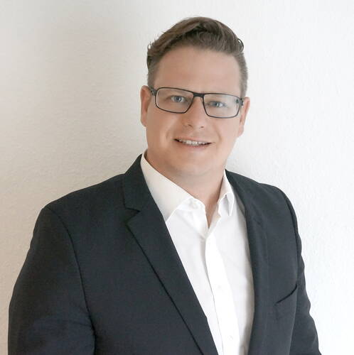 Mann mit Brille im dunklen Anzug und weißem Hemd lächelt vor einer weißen Wand in einem professionellen Porträt.