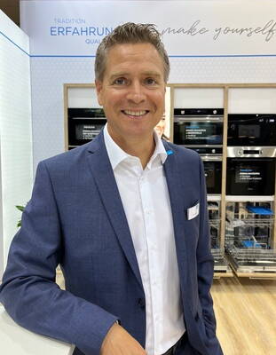 Lächelnder Mann im blauen Anzug steht in einem modernen Elektrogeräte-Showroom mit offenen Einbau-Spülmaschinen und Backöfen im Hintergrund.