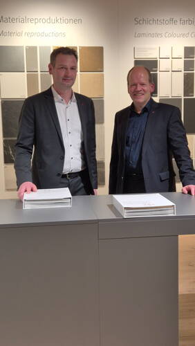 Zwei Männer im Anzug stehen in einer Ausstellung vor Wandtafeln mit Material- und Schichtstoffmustern und präsentieren jeweils einen Musterkatalog auf einem grauen Tresen.