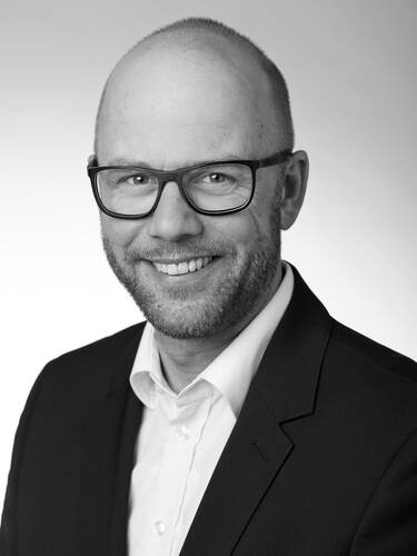 Lächelnder glatzköpfiger Mann mit Bart und Brille im Anzug vor neutralem Hintergrund