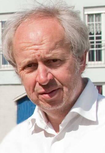 Älterer Mann mit lichtem, grauem Haar und weißem Hemd steht im Freien vor einem Gebäude mit Fenstern und blauer Tür.