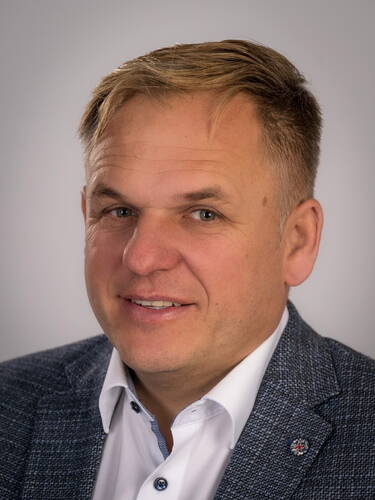 Mittelalterlicher Mann mit blondem Kurzhaarschnitt im grauen Sakko und weißem Hemd lächelt leicht vor neutral grauem Hintergrund
