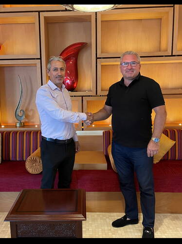 Zwei Männer in Businesskleidung geben sich vor einer dekorativen Holzregalwand mit roten und goldenen Elementen die Hand und besiegeln ein Treffen in einer Lounge-Umgebung