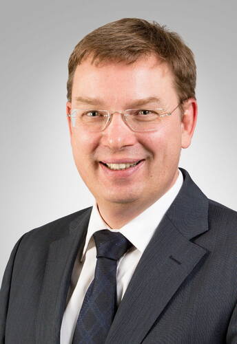 Lächelnder Mann mit kurzem braunem Haar, Brille und dunklem Anzug vor neutral grauem Hintergrund