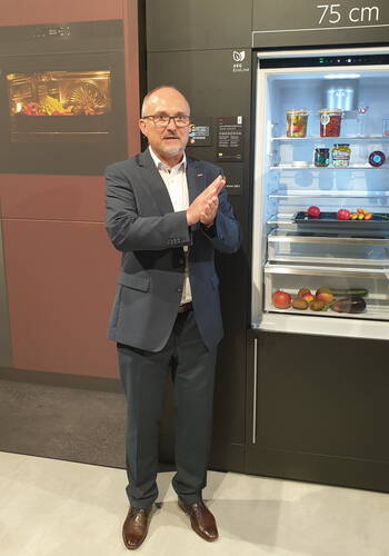 Mann im Anzug präsentiert einen geöffneten AEG-Kühlschrank neben einem modernen Backofen in einer Ausstellungsküche