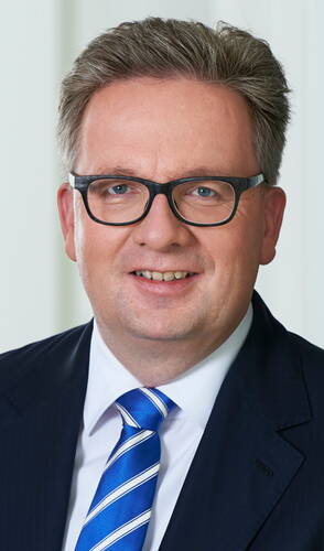 Mittelalterlicher Mann mit kurzem, grau meliertem Haar, schwarzer Brille und dunklem Anzug mit blau-weiß gestreifter Krawatte lächelt frontal in die Kamera vor hellem Hintergrund