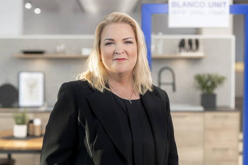 Selbstbewusste blonde Frau im schwarzen Blazer steht in einer modernen, hellen Küchenausstellung und blickt direkt in die Kamera