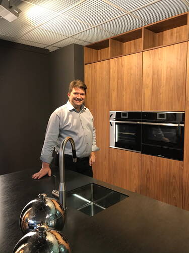 Mann lehnt lächelnd an einer modernen Kücheninsel mit schwarzer Arbeitsplatte, Edelstahlspüle und holzvertäfelter Einbauküche im Hintergrund