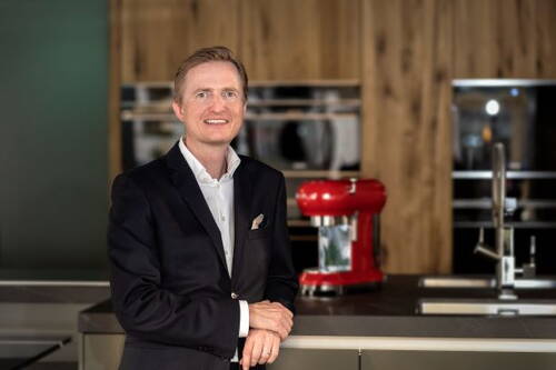 Mittelalter Mann im dunklen Anzug lächelt in einer modernen Küche und lehnt sich an eine Arbeitsplatte, im Hintergrund eine rote Küchenmaschine und Einbaugeräte vor Holzfronten