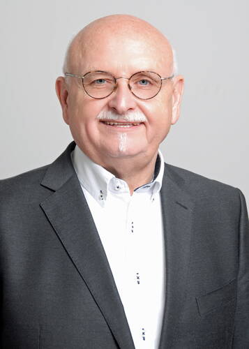 Älterer, glatzköpfiger Mann mit rundem Brillengestell, weißem Schnurrbart und kleinem Kinnbart lächelt in grauem Anzug und weißem Hemd vor neutralem Hintergrund.