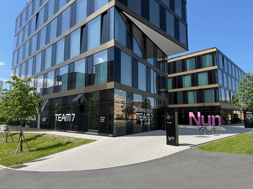 Moderne Glasbürogebäude mit TEAM7‑Showroom und großem pinken „Nun“-Schriftzug an einem sonnigen, begrünten Vorplatz