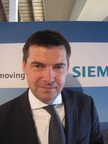 Mittelalterlicher Mann im dunklen Anzug und Krawatte steht vor einem Messestand mit teilweisem Siemens‑Schriftzug und blickt direkt in die Kamera