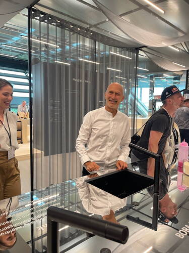 Lächelnder Mann in weißem Hemd steht hinter einer glänzenden, modernen Küchentheke mit schwarzer Spüle in einer hellen, futuristischen Ausstellungshalle