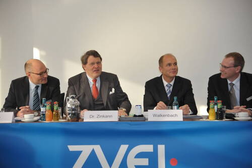 Vier Geschäftsleute im Anzug sitzen bei einer ZVEI-Pressekonferenz hinter einem blauen Podium mit Namensschildern, Getränken und Konferenzunterlagen.