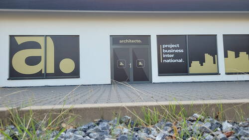 Eingang eines modernen Bürogebäudes mit architekto.-Beschriftung, großem „a.“-Logo und Fensterfolien mit der Aufschrift „project business international“ sowie stilisierter Stadtsilhouette im Vordergrund einer Pflasterfläche mit Kies und Gras.