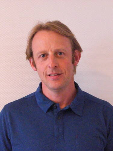 Mittelalterlicher Mann mit blondem Haar und blauem Poloshirt steht vor einer hellen strukturierten Wand und lächelt leicht in die Kamera.