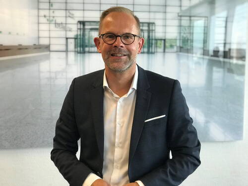 Mittelälterer Mann mit Brille im dunklen Anzug lächelt in moderner, lichtdurchfluteter Büro-Lobby in die Kamera