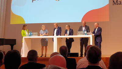 Podiumsdiskussion mit sechs Geschäftsleuten, die an einem hohen weißen Tisch auf einer Konferenzbühne vor sitzendem Publikum debattieren