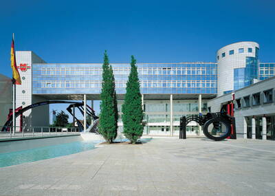 Moderner Bürokomplex mit verglaster Fassade, Wasserbecken, zwei hohen Nadelbäumen und schwarzer Metallskulptur im weitläufigen Innenhof unter blauem Himmel