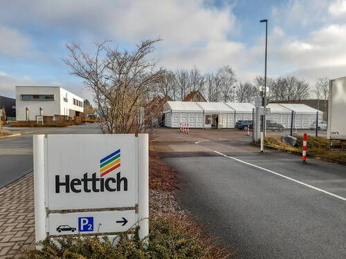 Einfahrt zum Hettich-Betriebsgelände mit Firmenschild im Vordergrund und weißen Hallenzelten sowie parkenden Autos im Hintergrund