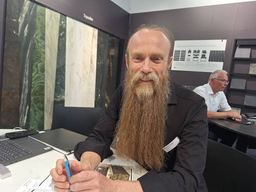 Mann mit sehr langem Vollbart sitzt lächelnd an einem Messetisch mit Unterlagen und Laptop, im Hintergrund Wand mit Steinmustern und ein weiterer arbeitender Mann