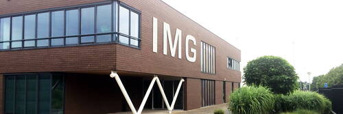 Modernes zweistöckiges Backsteingebäude mit großen Fenstern und weißen Stützpfeilern, an dessen Fassade die Buchstaben „IMG“ prangen, umgeben von gepflegten Grünanlagen.