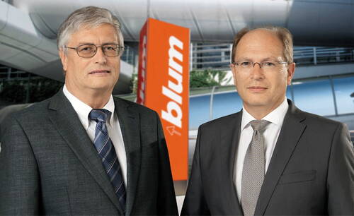 Zwei geschäftlich gekleidete Männer mit Brille stehen vor einem modernen Firmengebäude mit orangefarbenem „blum“-Firmenschild im Hintergrund