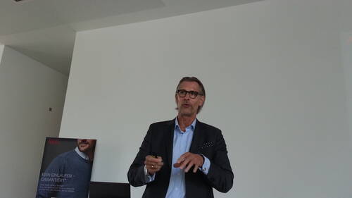 Mittelalterlicher Mann im Anzug hält einen Vortrag in einem modernen, hellen Konferenzraum und gestikuliert mit einem Präsentationsklicker in der Hand.