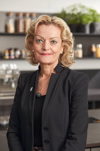 Elegante Geschäftsfrau mit blondem, gelocktem Haar und dunklem Blazer steht lächelnd in einer modernen Küche mit Gewürzregalen und Kräutertöpfen im Hintergrund.