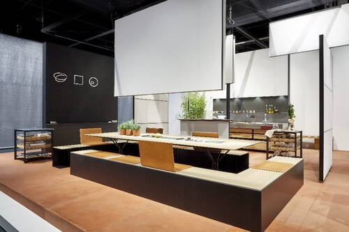 Modern gestalteter Messestand mit langem, niedrigem Holztisch, integrierten Sitzbänken, Pflanzenarrangements und minimalistischem Küchenbereich im Hintergrund.
