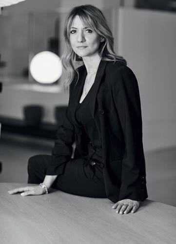 Elegante Frau im schwarzen Hosenanzug sitzt nachdenklich auf einer Tischkante in einem modernen, minimalistisch eingerichteten Raum in Schwarz-Weiß-Aufnahme
