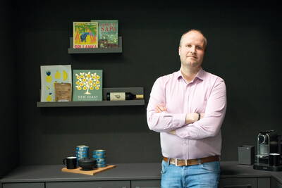 Mann mit verschränkten Armen in modernem Büroraum, neben dunkler Küchenzeile mit Kaffeemaschine, Konservendosen und dekorativen Regalen mit Kochbüchern und Weinflasche
