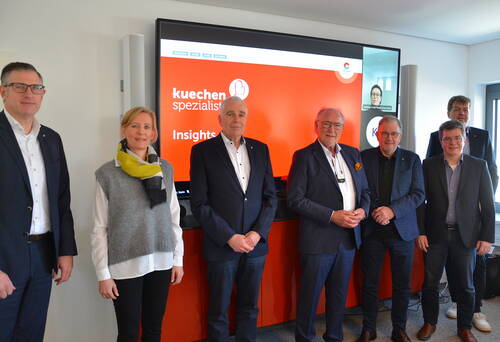 Mehrere Geschäftsleute stehen lächelnd in einem Konferenzraum vor einem großen Bildschirm mit rotem „kuechen spezialisten“-Hintergrund, während eine weitere Person per Videokonferenz zugeschaltet ist.