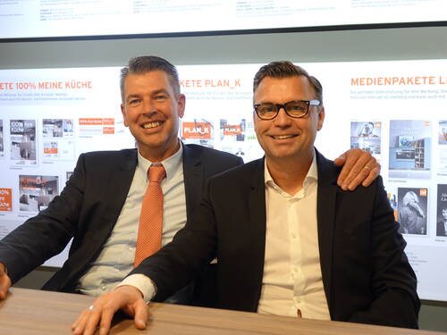 Zwei lächelnde Geschäftsmänner im Anzug sitzen nebeneinander an einem Tisch, einer legt freundschaftlich den Arm um den anderen, während hinter ihnen Präsentationsfolien zu Marketing- und Medienpaketen angezeigt werden.