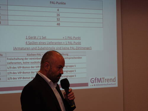 Mann im Anzug hält ein Mikrofon und präsentiert vor einer projizierten Folie mit PAL-Punkten und Bonusregelungen der Möbel-Einkaufsgruppe GfMTrend
