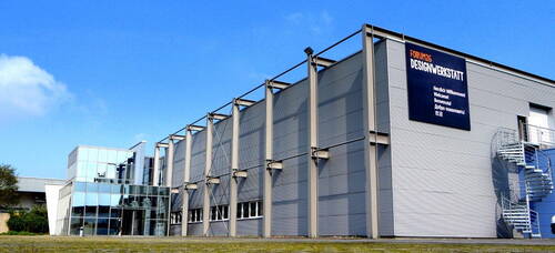 Modernes Industriegebäude „Forum 26 Designwerkstatt“ mit verglaster Eingangsfassade, grauen Metallpaneelen und außenliegender Wendeltreppe unter blauem Himmel