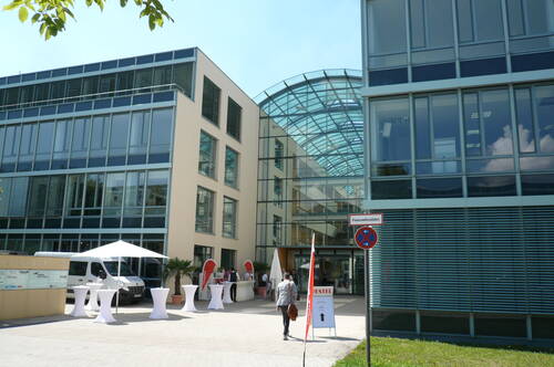 Modernes Bürogebäude mit verglastem Eingangsbereich, Stehtischen und Infostand bei einer Veranstaltung, während eine Person mit Aktentasche auf den Eingang zugeht.