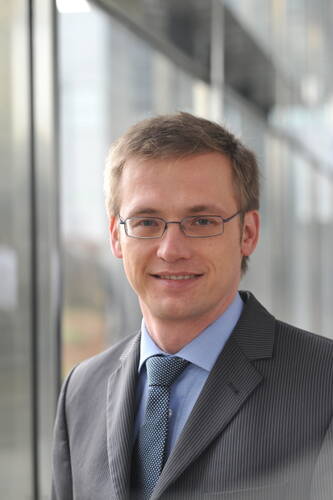 Junger Mann im Anzug und mit Brille lächelt in einem modernen Bürogebäude in die Kamera