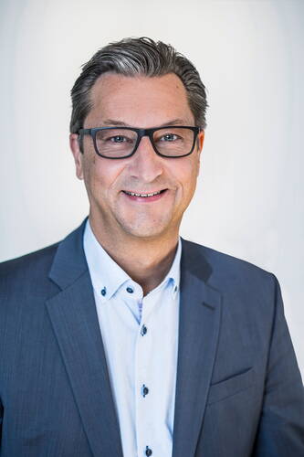 Mittelälterer Mann mit Brille und graumeliertem Haar lächelt in die Kamera und trägt ein blaues Sakko mit hellblauem Hemd vor neutralem Hintergrund