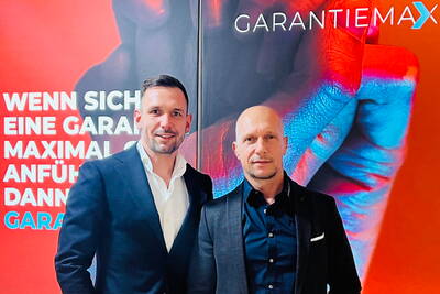 Zwei geschäftlich gekleidete Männer posieren lächelnd vor einer rot-blauen Werbewand mit dem Schriftzug „GARANTIEMAX“ und einem großformatigen Motiv einer farbig beleuchteten Hand.