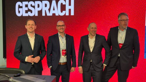 Vier Geschäftsleute in dunklen Anzügen stehen lächelnd vor einer großen roten Leinwand mit dem Wort „GESPRÄCH“ im Hintergrund während einer Konferenz.