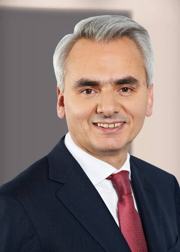 Älterer Mann mit grauen Haaren im dunklen Anzug und roter Krawatte lächelt direkt in die Kamera vor neutralem Hintergrund