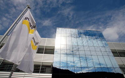Moderne Unternehmensfassade mit spiegelnden Glasfronten, in denen sich Himmel und Wolken reflektieren, daneben zwei im Wind wehende weiße Firmenflaggen.