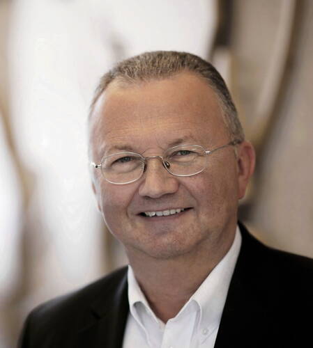 Älterer Mann mit Brille und kurzem grauem Haar lächelt freundlich in die Kamera und trägt ein weißes Hemd unter einem dunklen Sakko vor unscharfem Hintergrund.