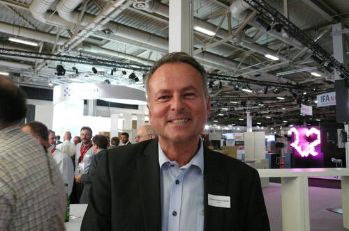 Lächelnder Mann im Anzug mit Namensschild steht in einer belebten Messehalle mit modernen Messeständen und Beleuchtung im Hintergrund